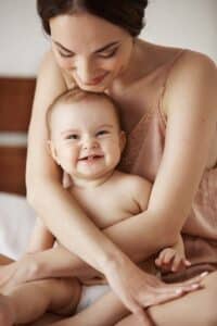 Young beautiful happy mom in sleepwear and her newborn baby sitting on bed smiling playing together at home. Copy space.