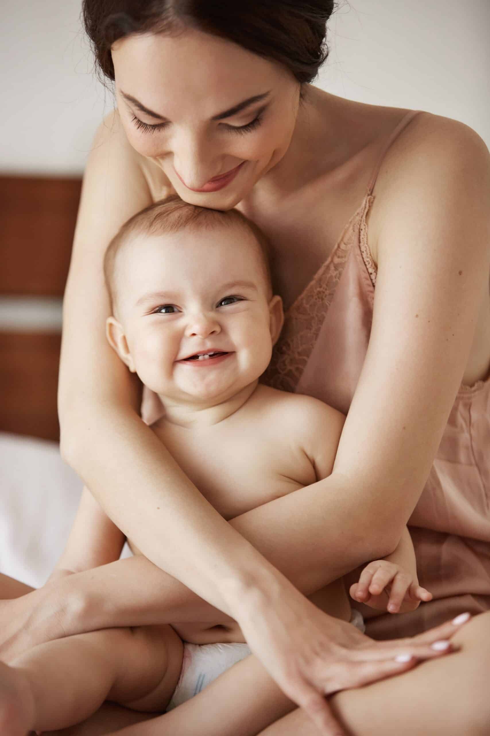 Young beautiful happy mom in sleepwear and her newborn baby sitting on bed smiling playing together at home. Copy space.