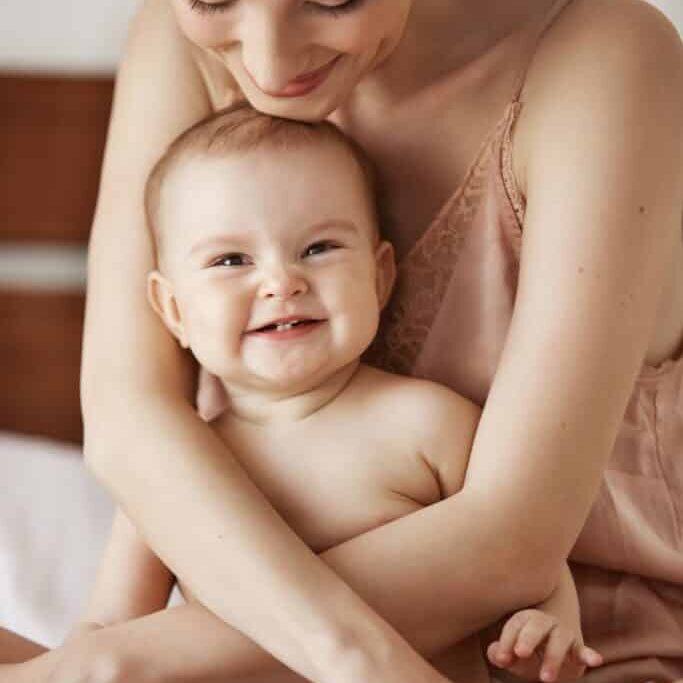 Young beautiful happy mom in sleepwear and her newborn baby sitting on bed smiling playing together at home. Copy space.